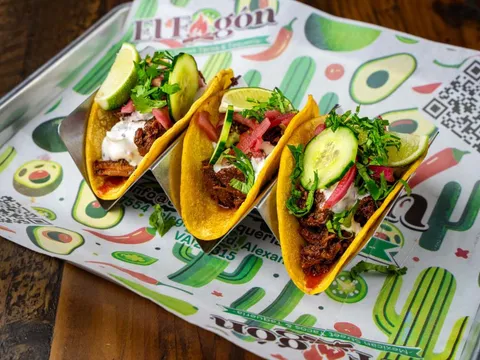 Three El Fogón Taquería street tacos on a branded paper-lined metal tray, topped with cilantro, cucumber and pickled onion, with lime wedges