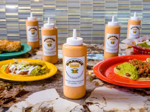 Bottles of La Victoria Taqueria's signature Orange Sauce lined up on a marble counter beside plates of tacos